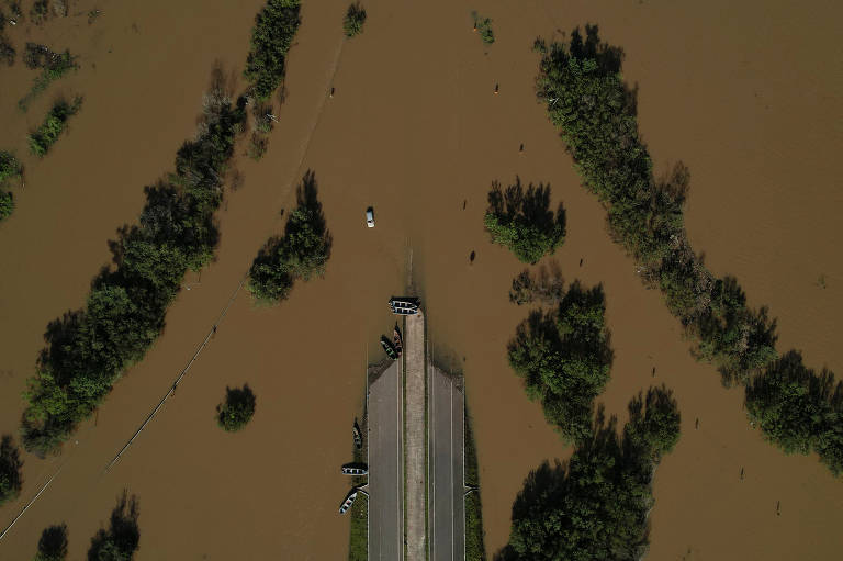 Uma vista aérea mostra uma estrada que acaba em uma inundação de água marrom, na qual aparecem umas árvores.