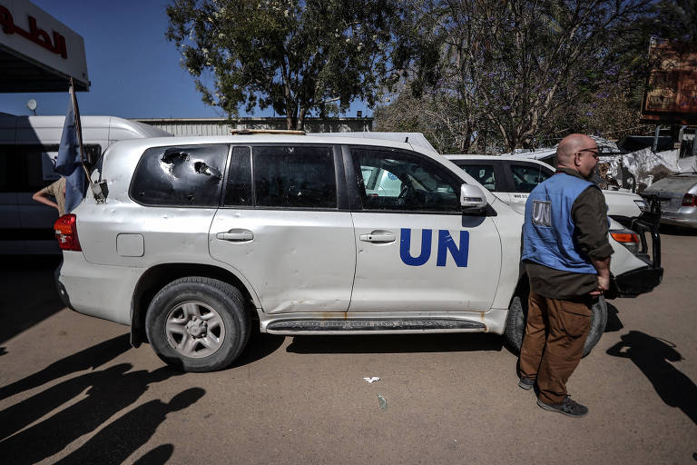 Homem com colete da ONU está parado em frente a um carro branco da organização, que tem os vidros de trás quebrados após veículo ser alvejado