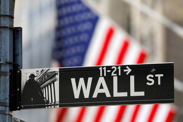 Placa indica uma rua de Wall Street, desfocada ao fundo está uma bandeira dos Estados Unidos