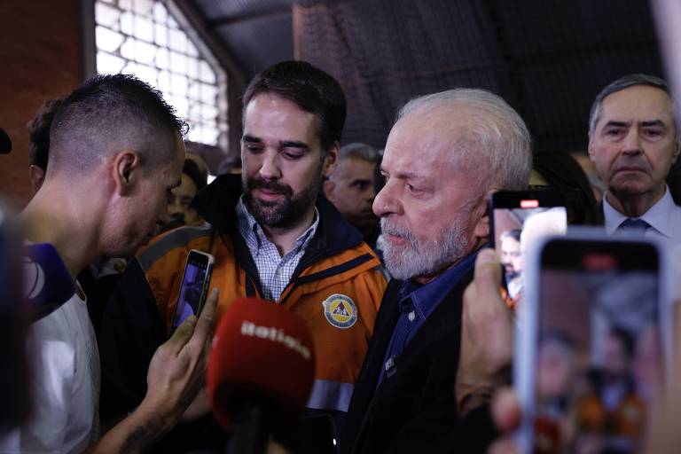Ao centro, governador Eduardo Leite, de abrigo cor de laranja, junto ao presidente Lula, de cabelo e barba brancos; à esquerda, uma jovem com um celular na mão direita e, ao fundo, o ministro do STF Luís Roberto Barroso, de camisa branca