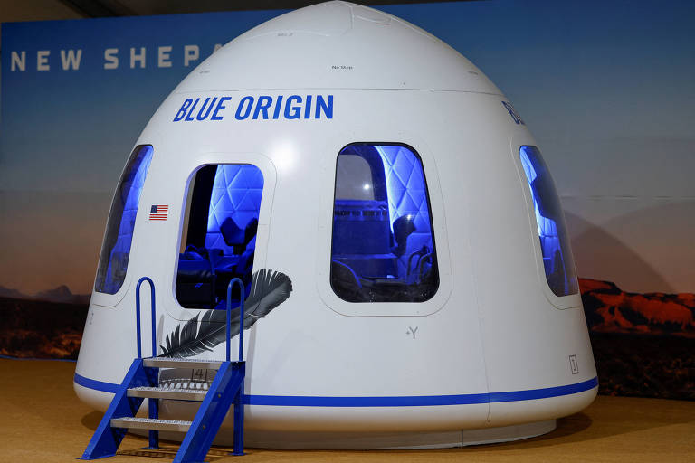A imagem mostra a cápsula de tripulação da Blue Origin, conhecida como New Shepard, projetada para levar turistas ao espaço. A cápsula branca com detalhes em azul possui grandes janelas para que os passageiros possam ver o espaço. Uma escada com corrimão azul permite o acesso ao interior da cápsula, que está estacionada em um ambiente que simula uma base de lançamento.
