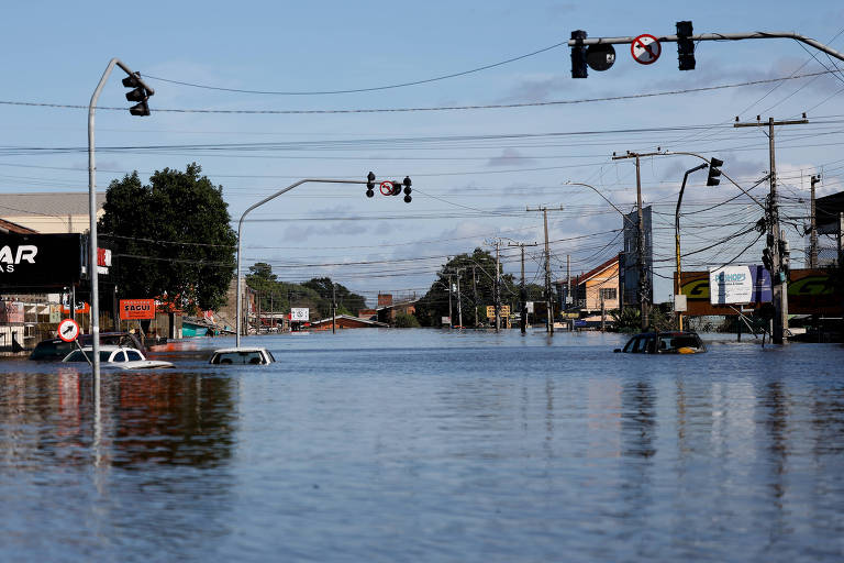 Rua em Canoas (RS) ainda está alagada mais de três semanas após início das enchentes que atingiram o estado. Imagem mostra carros com água até a altura do vidro frontal do veículo