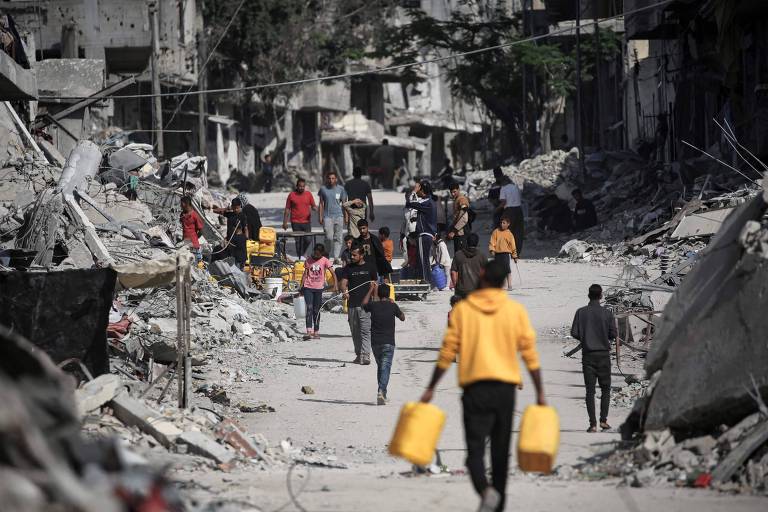 Homem de blusa amarela e calça preta segura dois potes amarelos, um em cada mão, enquanto caminha em Gaza, que está em escombros