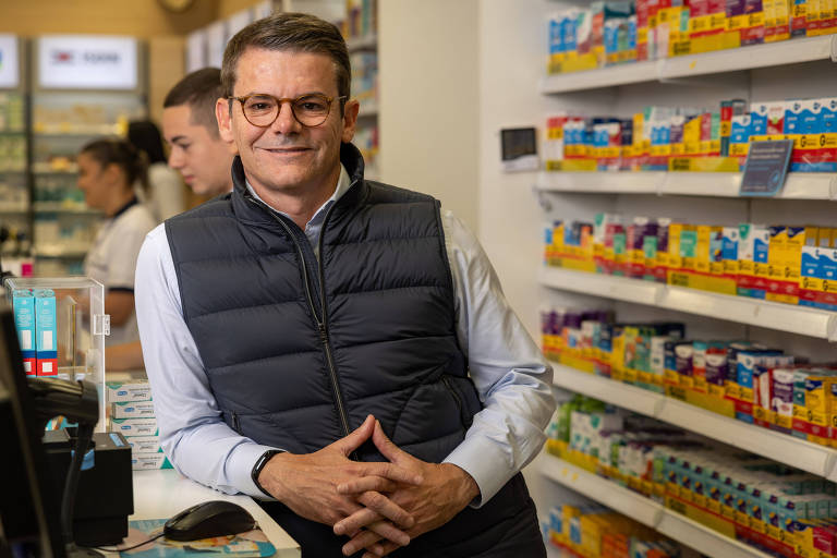Homem branco de óculos e colete preto em balcão de farmácia