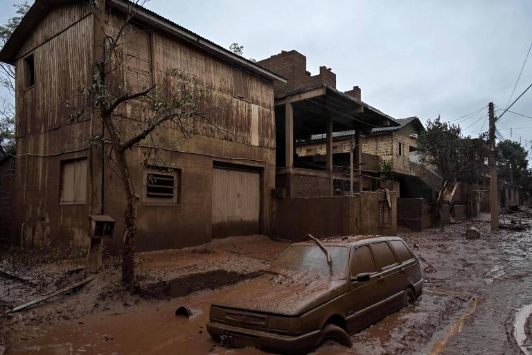 Rua de bairro residêncial completamente coberto por lama. Uma casa de dois andares com marcas de lama até o telhado. Em frente à casa, um carro completamente coberto por lama.