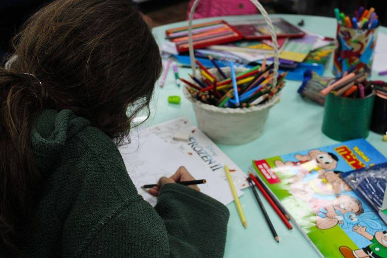 Na foto, uma criança de costas está pintando uma folha de papel com lápis de cor