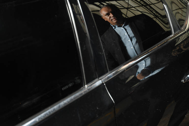 A imagem mostra o reflexo de um homem, vestido com terno sem gravata e camisa azul claro, no vidro de um carro preto. A expressão em seu rosto é séria