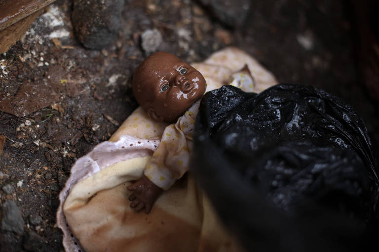 Um brinquedo quebrado, possivelmente uma boneca, em meio a detritos e um saco plástico preto