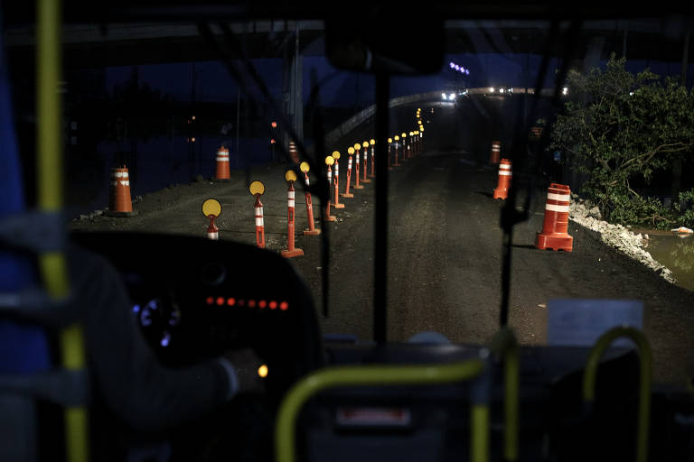 Vista de dentro de um ônibus, uma estrada com sinalização temporária é vista ilumidada pelos faróis do veículo.