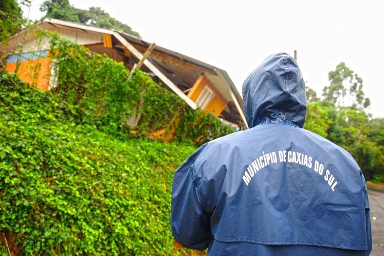 Funcionário municipal de Caxias do Sul, vestindo capa de chuva azul com o nome do município estampado nas costas, observa construção parcialmente destruída