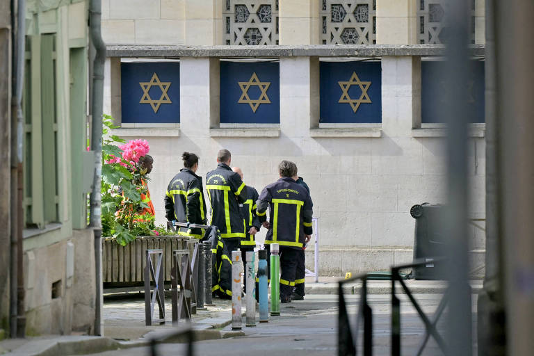 Bombeiros uniformizados em pé, em frente a uma sinagoga, identificável pelas estrelas de Davi na lateral do templo