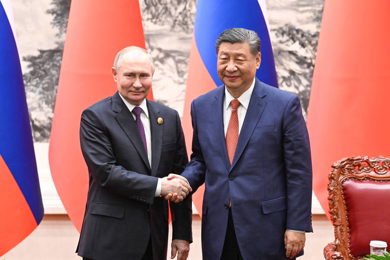 Vladimir Putin troca aperto de mãos com Xi Jinping enquanto ambos sorriem para fotógrafos. O líder russo está usando um terno preto, ao lado esquerdo da foto. O dirigente chinês está ao lado direito, usando um terno azul. Ao fundo podemos ver bandeiras dos dois países intercaladas.