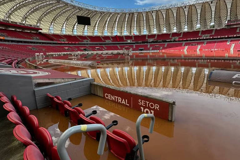 Na foto, um estádio com uma cobertura branca e cadeiras vermelhas está parcialmente alagado. Onde estaria o gramado, há apenas água marrom