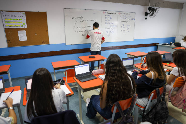 Um professor está de costas para a câmera, escrevendo em um quadro branco, enquanto os alunos, sentados em carteiras com laptops abertos, prestam atenção à lição