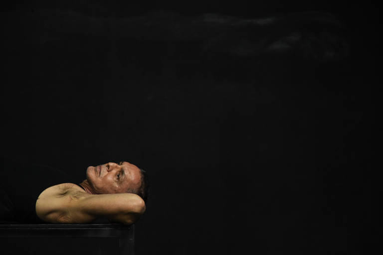 Homem de regata preta deitado, fundo escuro