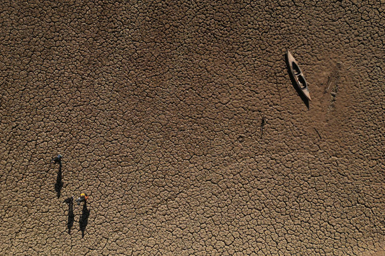 A fotografia foi tirada com drone e mostra, do alto, uma canoa e duas pessoas em solo marrom, muito seco, com a terra cheia de rachaduras