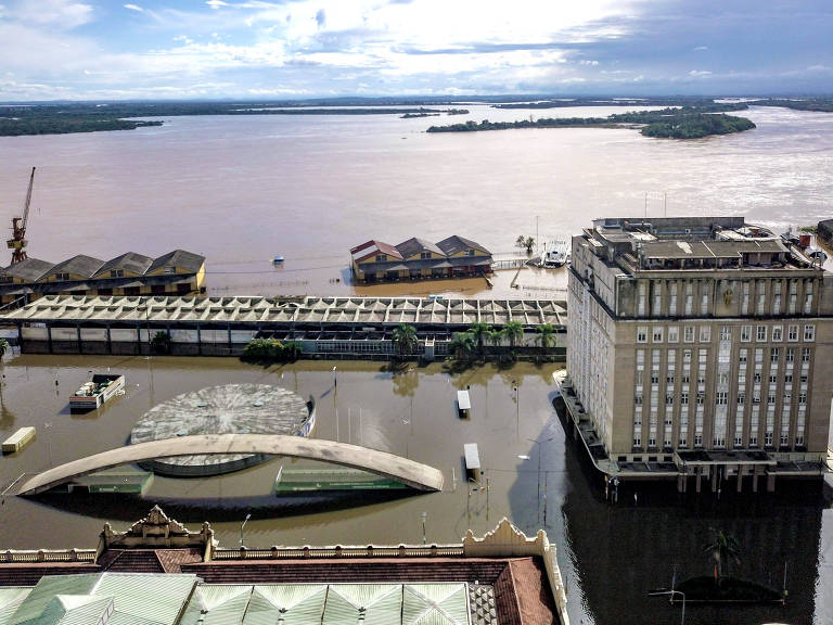 A imagem captura a vista aérea da cidade de Porto Alegre com o lago Guaíba ao fundo e as águas entre os prédios, no primeiro plano