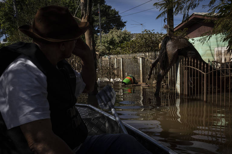 Um homem com chapéu de aba larga e colete observa atentamente enquanto navega em uma pequena embarcação por uma rua inundada
