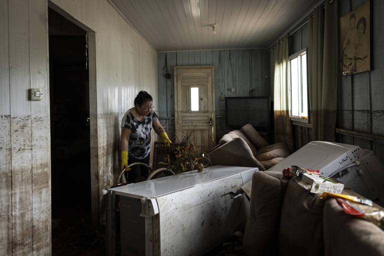 Uma mulher parece estar limpando uma sala de estar que foi devastada por uma inundação. Móveis e eletrodomésticos, incluindo um sofá e uma geladeira, estão cobertos de lama e detritos