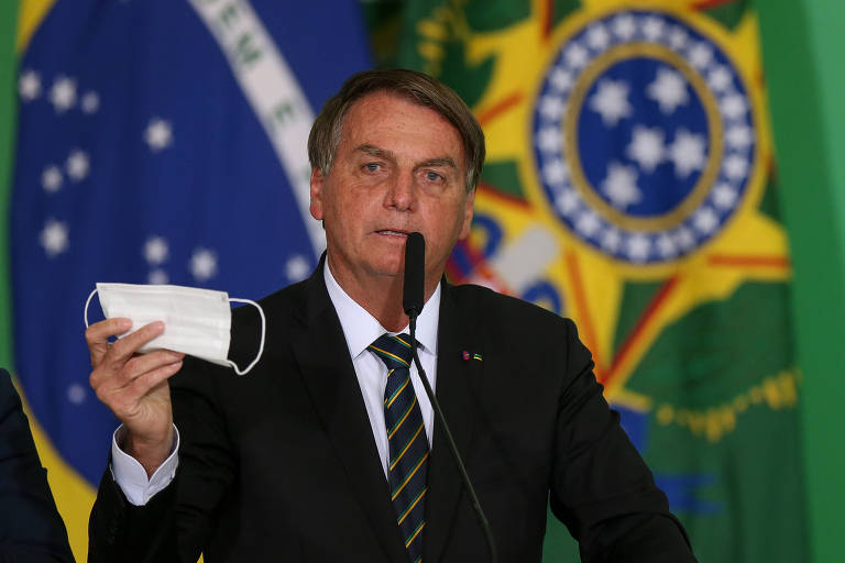 Bolsonaro, um homem branco, cabelos castanhos e terno, está diante de um microfone de mesa, segurando uma máscara com a mão esquerda. Ao fundo, duas bandeiras, a do Brasil e a presidencial