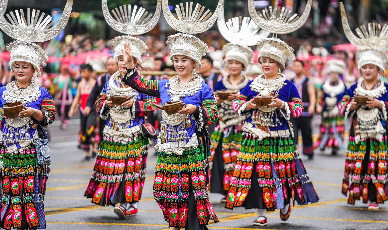 A imagem mostra um grupo de mulheres vestidas com trajes tradicionais coloridos e elaborados, desfilando em uma rua. Elas usam grandes chapéus prateados em forma de meia-lua com detalhes ornamentais e colares de contas brancas. As roupas são predominantemente azuis e pretas, com saias decoradas com padrões florais multicoloridos