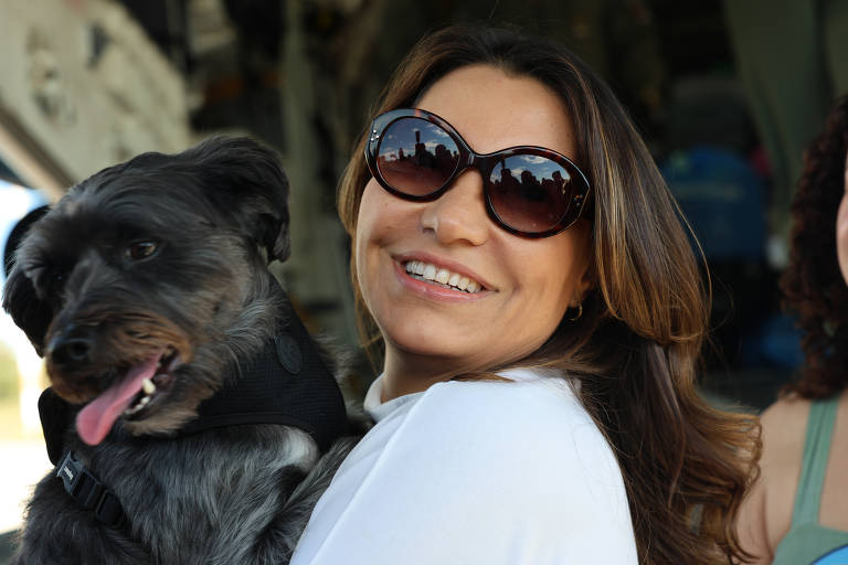 A imagem mostra uma mulher sorridente segurando um cão de pequeno porte. Ela usa óculos de sol grandes com lentes refletivas, através das quais se pode ver o reflexo de algumas pessoas e estruturas. O cão tem pelo curto, preto e cinza, e está usando um arnês preto