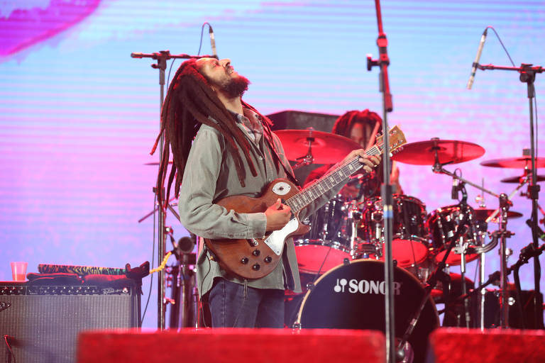 Julian Marley, cantor de reggae, está no palco, trajado de blusa cinza e calça jeans. Ele olha para o céu enquanto toca sua guitarra. Ao fundo está o baterista da banda, que aparece atrás do instrumento