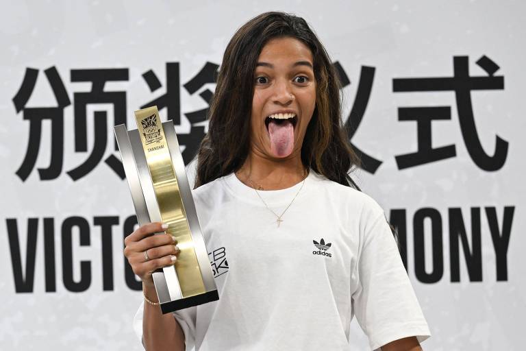 Uma jovem está posando para a foto, segurando um troféu em uma mão e mostrando a língua. Ela usa uma camiseta branca e tem cabelo longo e solto. Ao fundo, há um painel com texto em caracteres asiáticos e a palavra 'VICTORY CEREMONY'.