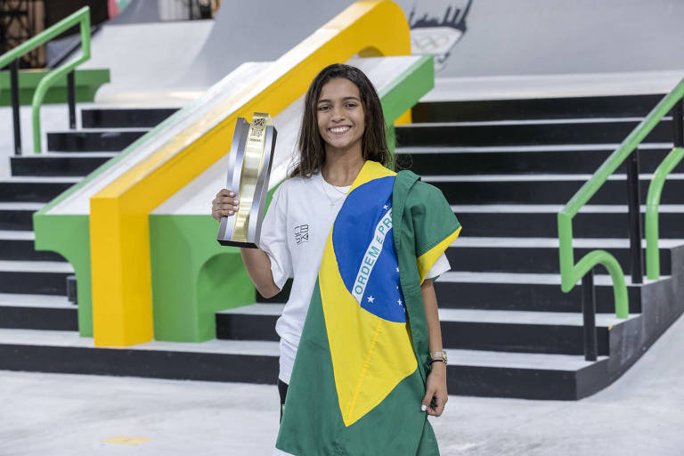 A skatista Rayssa Leal, enrolada em ua bandeira do Brasil, sorri e exibe um troféu
