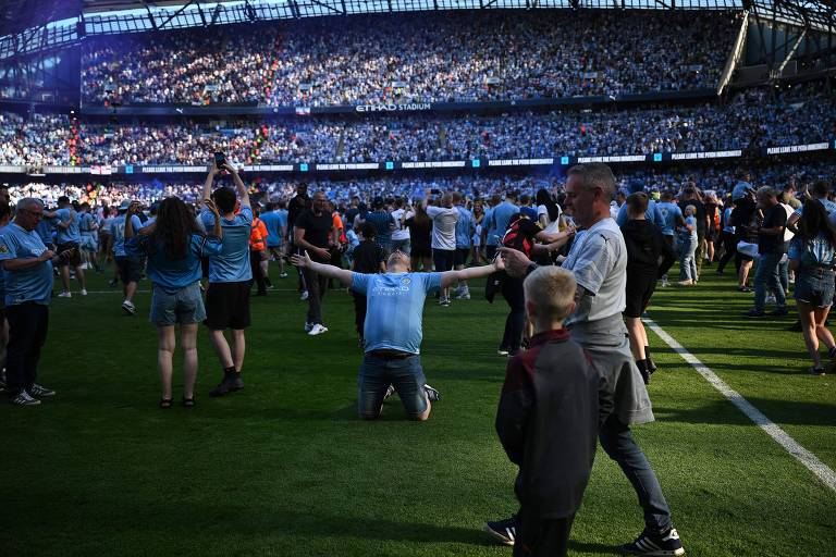 De joelhos, com os braços abertos e vestindo a camisa azul celeste do time, torcedor do Manchester City celebra a conquista da Premier League no gramado do estádio Etihad, invadido por simpatizantes da equipe