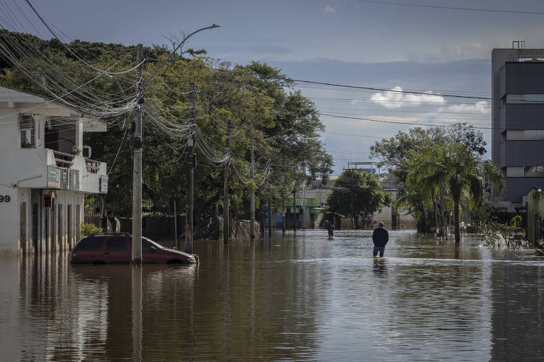 Homem caminha em alagamento em Eldorado do Sul, passando ao lado de um carro parcialmente coberto pelas águas