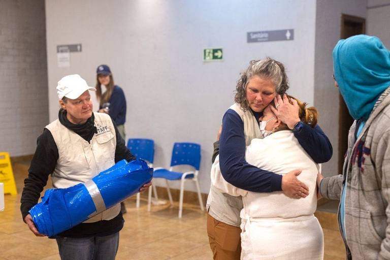 Em um abrigo, uma voluntária abraça uma senhora que usa máscara de proteçaõ e está chorando