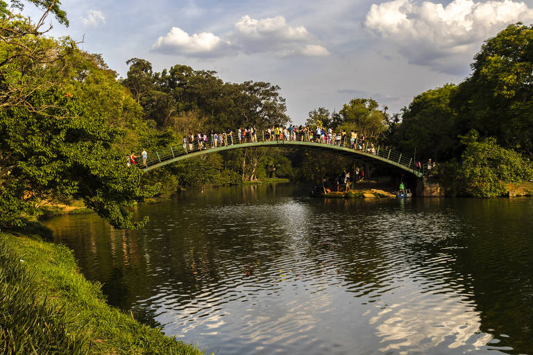 Paulistanos curtem dia de sol em ponte sobre o lago do Parque do Ibirapuera; em primeiro plano, as águas do lago e, ao fundo, muitas árvores