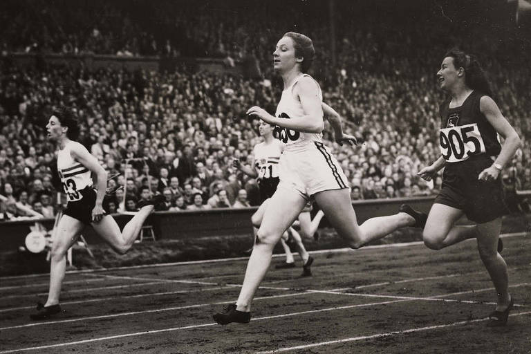 Foto em preto e branco mostra mulheres em competição de atletismo. Ao centro, uma mulher vestida com camiseta e short corre.