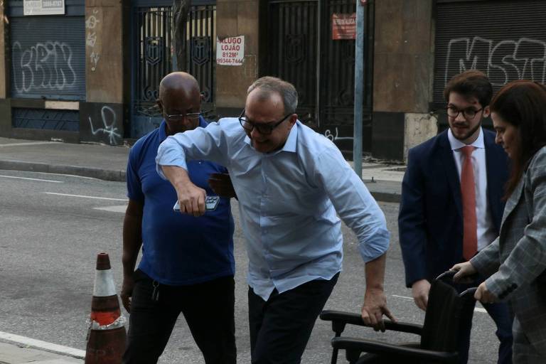 Na imagem, Sérgio Cabral, de camisa azul clara e óculos, é escoltado apressadamente por um homem de camisa azul escura, enquanto outro homem e uma mulher em trajes formais os seguem de perto
