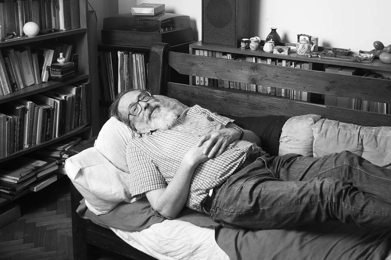 Homem branco de barba grisalha e óculos deitado em um banco com as pernas esticadas dentro de uma sala com uma estante cheia de livros ao fundo