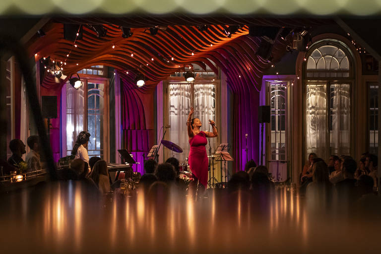 No palco da Casa de Francisca, a cantora Adriana Moreira usa um vestido vermelho. A artista está em pé, no centro da foto, em um palco iluminado por luzes roxas e âmbar. O teto do local tem testo de madeira curvado e janelas altas.