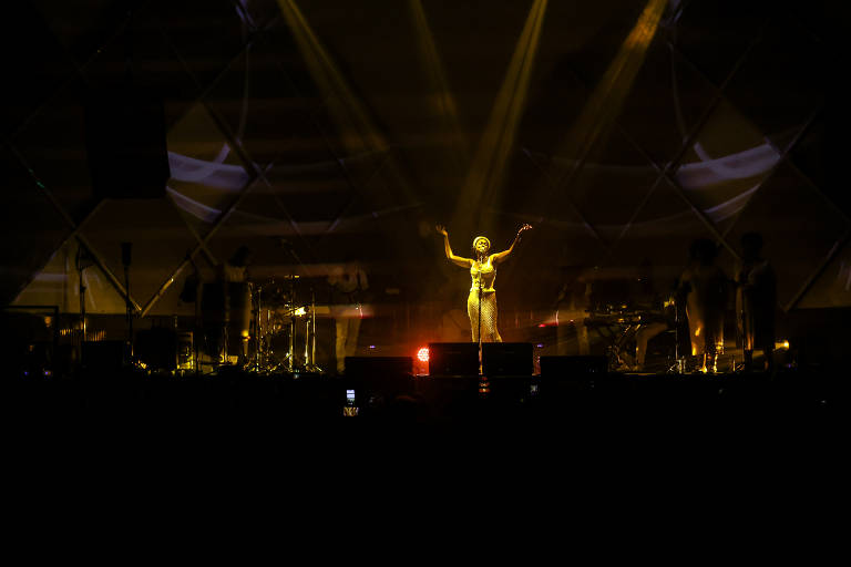 No palco no Cine Joia, na parte central, uma artista é iluminado por luzes amarelas, destacando-se contra um fundo escuro. Ela está com os braços abertos enquanto canta