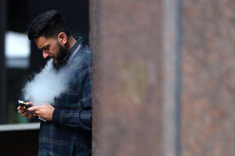 A imagem mostra um homem com barba, vestindo uma camisa xadrez, usando um vape enquanto olha para o celular. Ele está encostado em uma parede de pedra, e uma nuvem de vapor é visível ao seu redor.