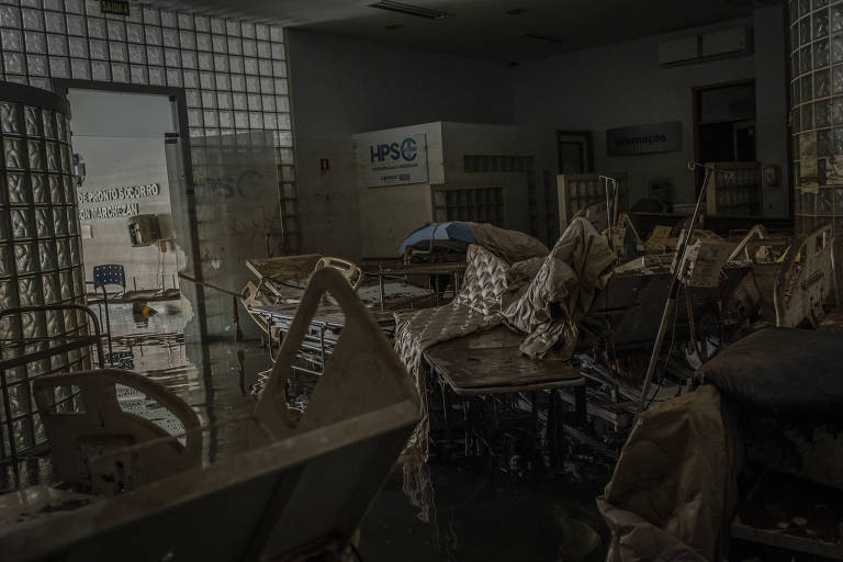 A imagem mostra o interior de um hospital abandonado e em ruínas, com cadeiras de rodas e camas hospitalares desordenadas e cobertas de detritos. O chão está inundado de água