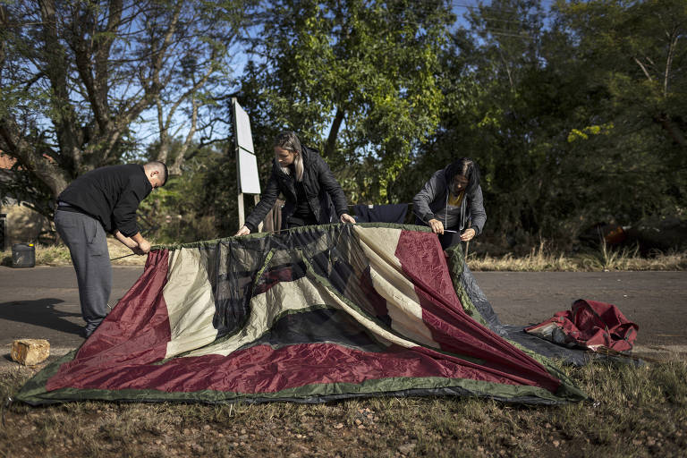 Três pessoas trabalham juntas para montar uma barraca em um ambiente ao ar livre. Os três estendem e ajustam o tecido da barraca sobre o chão coberto de folhas secas.
