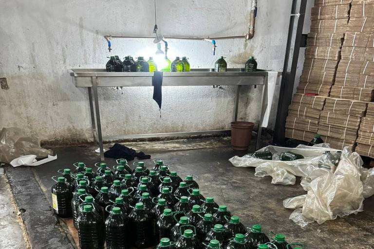 A imagem mostra um espaço de armazenamento com várias garrafas de azeite de oliva verde-escuras alinhadas no chão de concreto. Acima, há uma pia de metal com algumas garrafas, e ao fundo, pilhas de caixas de papelão