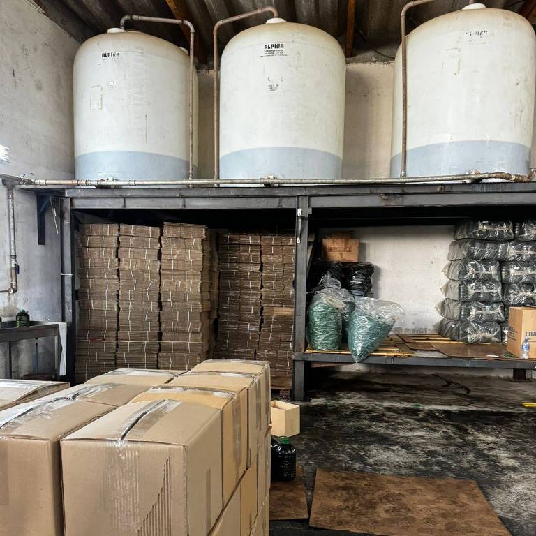A imagem mostra um armazém industrial com três grandes tanques de armazenamento brancos posicionados sobre uma estrutura de metal no alto. Abaixo, há pilhas de caixas de papelão e sacos empilhados