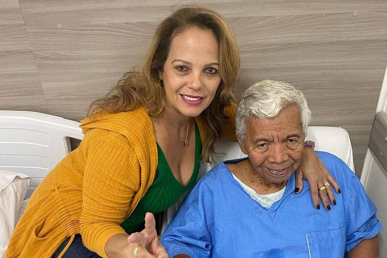 Janilda Nogueira, mulher sorridente com um cardigã amarelo e vestido verde está sentada ao lado do marido Roque, homem mais velho, que está vestindo uma bata de hospital azul e coberto com um cobertor. A mulher fazendo um gesto de paz com a mão.