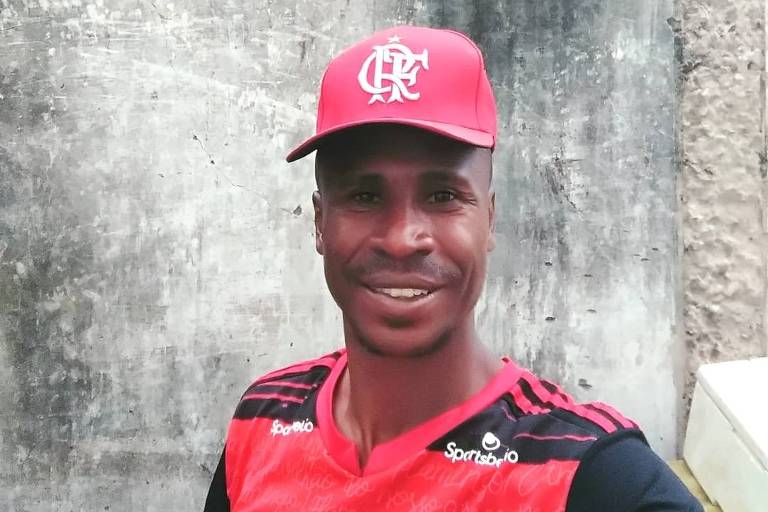 Um homem sorridente posa para a foto vestindo uma camisa e boné vermelhos do Flamengo, um famoso time de futebol brasileiro. Ele está em frente a uma parede de concreto desgastada, o que contrasta com as cores vibrantes de seu traje esportivo.