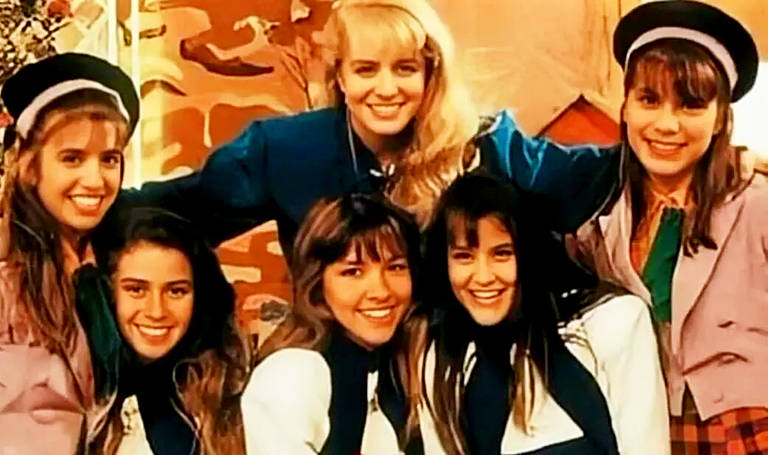 Um grupo de seis jovens mulheres sorri para a câmera, exibindo a moda e o estilo dos anos 90. Elas estão vestidas com roupas casuais que incluem jaquetas, camisas e acessórios como lenços e boinas, sugerindo uma atmosfera descontraída e amigável