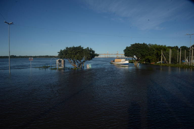 Uma paisagem inundada sob um céu claro, com árvores parcialmente submersas e um barco ancorado, sugerindo uma cena de calma após uma enchente
