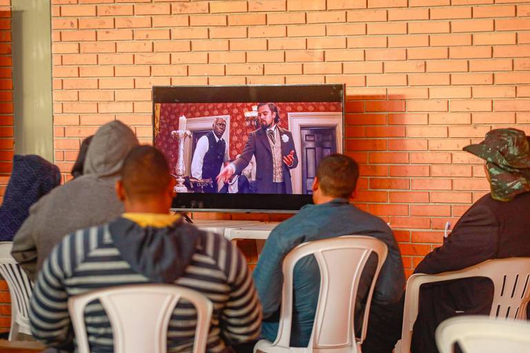 Na foto, cinco pessoas sentadas, de costas para a câmera, assistem o filme 'Django Livre' em uma televisão