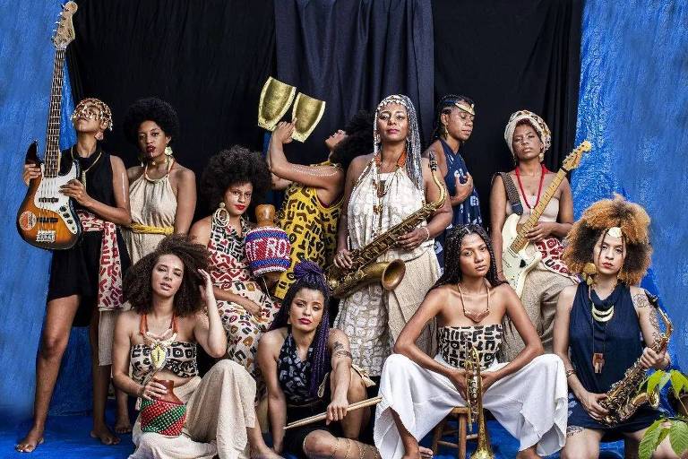 Orquestra Funmilayo Afrobeat, grupo composto por 11 mulheres negras, que posam em frente a um fundo azul. Cada uma segura um instrumento musical, como trompete, baixo e percussão