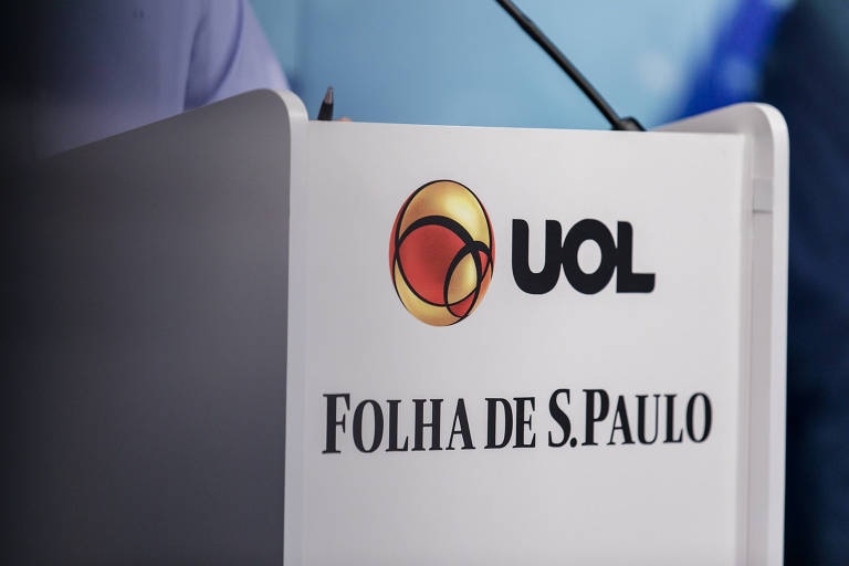 A imagem mostra um close-up de um suporte de microfone com os logotipos de UOL e Folha de S.Paulo, em debate das eleições de 2020